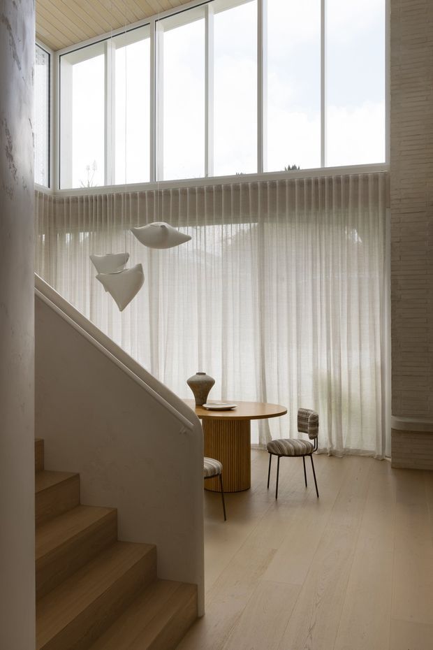 A softly lit, double-height interior with a timber staircase, pale timber flooring and ceiling, full-height corner glazing, sheer curtains, and a small round timber dining table with upholstered chairs. Sculptural pendant lights and a simple ceramic vessel add restrained decorative detail. The space has a warm, calm, minimalist character with strong vertical glazing and natural materials.