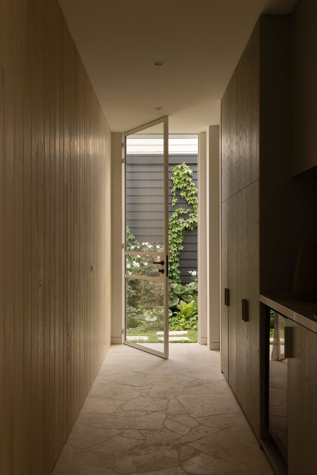 A narrow, warmly lit residential hallway leading through an open glazed door to a small landscaped courtyard. The corridor has full-height vertical timber wall panelling on the left, timber cabinetry or storage joinery on the right, recessed ceiling lights, and an irregular natural stone or flagstone floor. The open slender-framed door provides a view of dark horizontal exterior cladding, climbing greenery, and garden planting beyond.