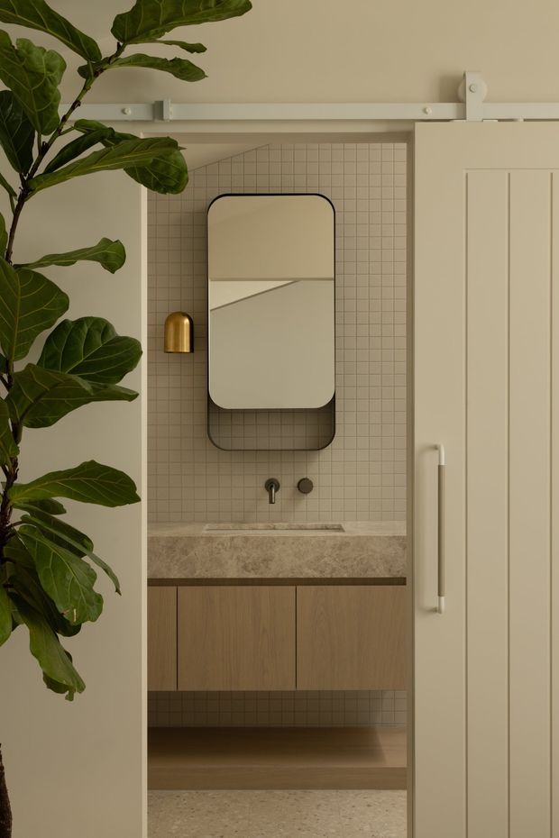 A warm, refined bathroom vanity nook viewed through a partially open cream sliding barn door. The space features small square beige wall tiles, a rounded rectangular black-framed mirror, brushed brass wall light, dark metal tapware, a textured stone or concrete benchtop, and vertical-grain timber cabinetry with an open shelf. A large leafy indoor plant frames the left side, while the white barn-door track and paneled door are visible above and to the right.