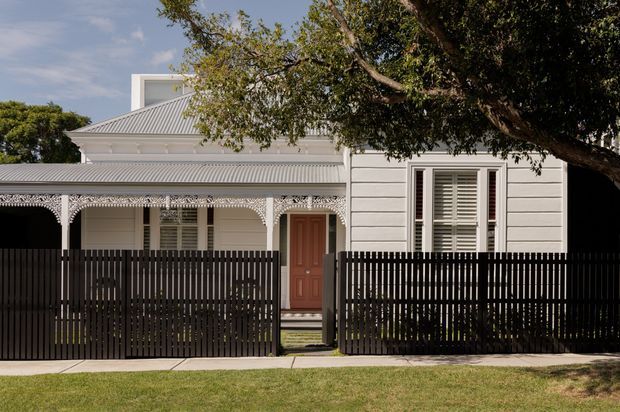Front exterior of a traditional single-storey home with white horizontal weatherboard cladding, a corrugated metal roof, decorative lacework verandah, timber shutters, and a warm timber entry door. A dark vertical-slat fence and mature tree frame the street-facing facade.