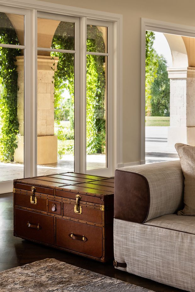 A refined sitting room corner with tall, white-trimmed multi-pane windows and a glazed door opening to a sunny covered exterior arcade. The room features a large upholstered sofa in a neutral woven fabric, a vintage brown leather-and-wood trunk used as a coffee table, dark timber flooring, and a textured neutral rug. The architecture and furnishings have a traditional, warm, understated character.