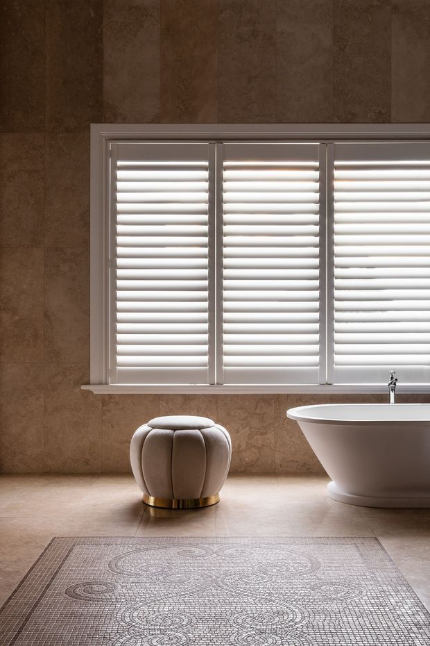 A refined bathroom interior featuring warm brown stone-look wall and floor finishes, a large white plantation-shuttered window, a freestanding white bathtub, and a rounded upholstered stool with a brass-toned base. A patterned area rug adds subtle texture in the foreground.