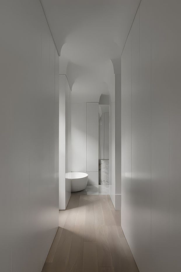A narrow, softly lit white hallway leading toward a minimalist bathroom area. The corridor has smooth white walls and integrated doors or cabinetry, a pale timber floor, and sculptural arched ceiling transitions. A freestanding white bathtub is visible at the end, with pale stone or tile surfaces beyond. The composition is monochromatic, refined, and highly minimal.
