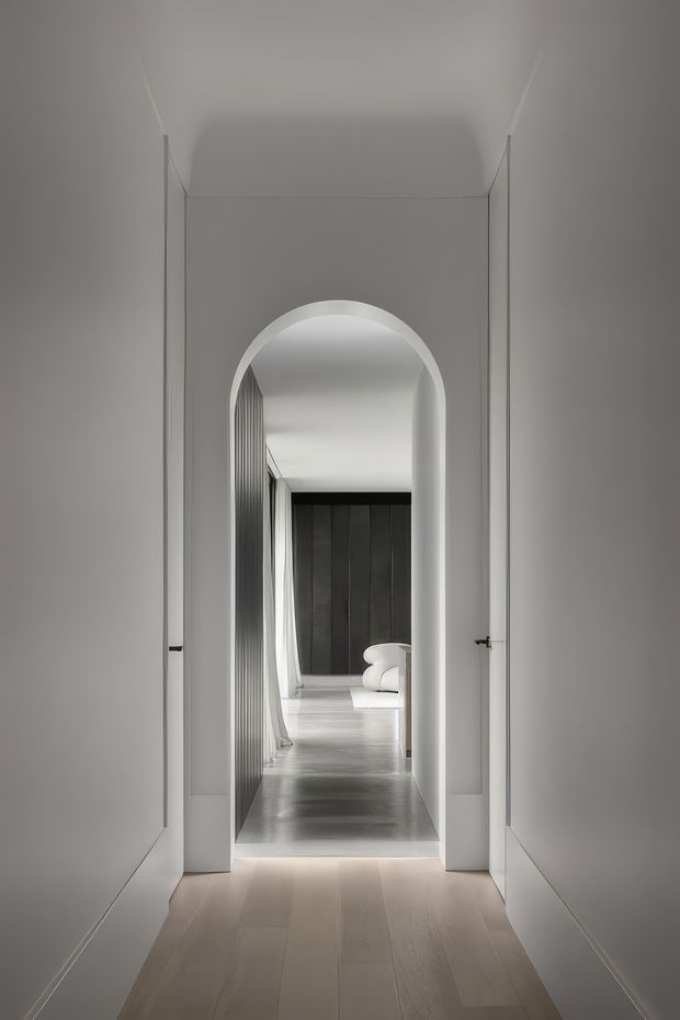 A minimalist interior hallway viewed through open double doors, centred on a tall rounded archway leading into a secondary living area. The corridor has smooth pale walls, simple linear trims, light timber flooring in the foreground, and a darker polished floor beyond the arch. A dark vertical-panel feature wall and part of a curved white sofa are visible in the distant room. The composition is symmetrical, restrained, and monochromatic with soft natural light.