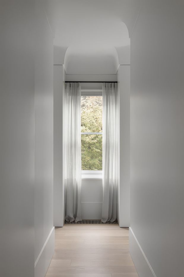 A narrow, minimalist interior passage or alcove leading to a tall window. The space has smooth white walls, a sculpted curved ceiling detail above the window, sheer white curtains, pale timber flooring, and simple white skirting boards. Soft daylight filters through the window, creating a calm and understated atmosphere.