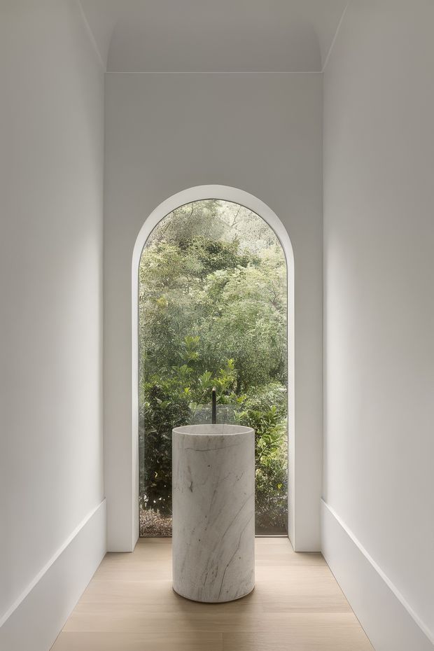 A narrow, serene interior corridor with smooth white walls and a light timber floor, terminating in a tall arched glazed opening overlooking dense greenery. A sculptural cylindrical stone basin or pedestal sits centrally in front of the window, creating a minimalist focal point. The space has clean geometric detailing, soft natural light, and a calm, gallery-like atmosphere.