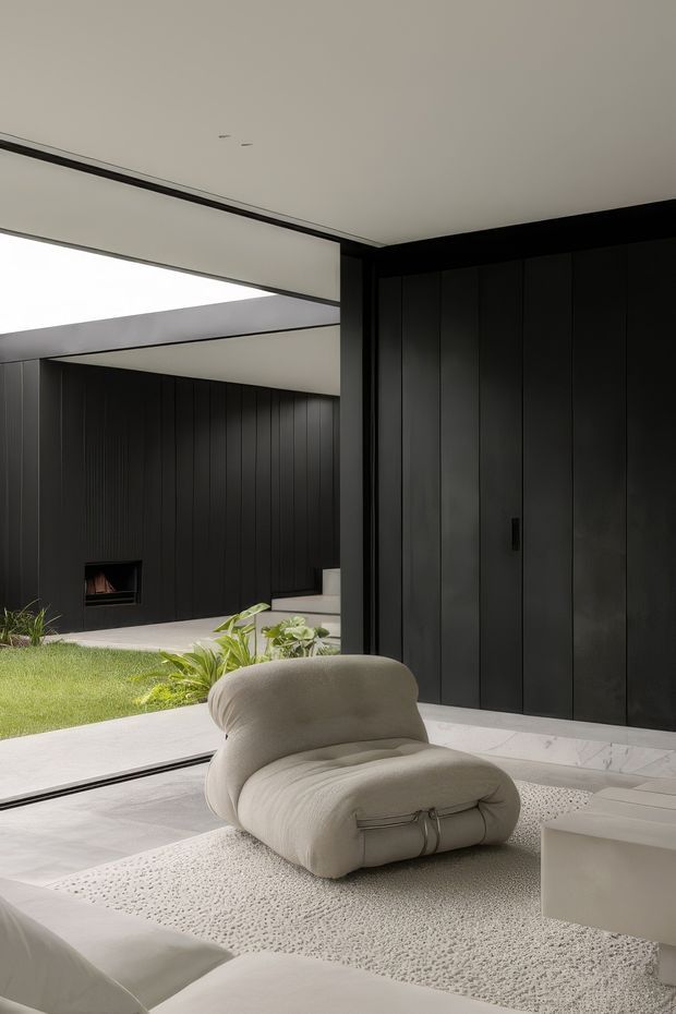 A refined contemporary interior centered on a sculptural cream lounge chair and pale textured rug. The space features dramatic full-height black vertical-panel joinery, pale stone or concrete flooring, a low white coffee table, and expansive glazing opening toward a landscaped courtyard. A dark feature wall with a small built-in fireplace is visible beyond the seating area, while concealed linear ceiling lighting and crisp horizontal architectural planes create a minimalist, gallery-like atmosphere.