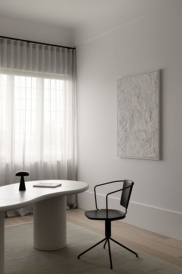A minimalist, monochromatic home office or study corner with a sculptural white oval desk, black task lamp, black modern armchair, sheer floor-to-ceiling curtains, pale timber flooring, and a framed abstract artwork. The room is softly lit by filtered daylight and has a calm, refined atmosphere.