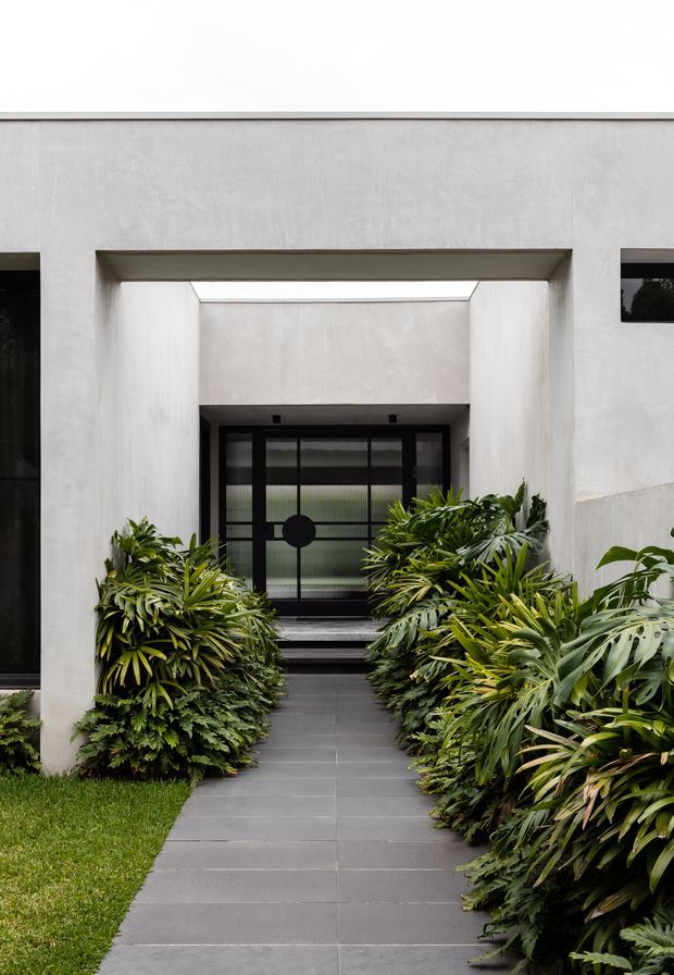 Front entry to a minimalist modern residence, framed by a deep rectangular concrete or rendered portal. A dark tiled pathway leads through lush tropical planting to a black-framed glazed entry door, with a skylit or open-air recess above the entrance.