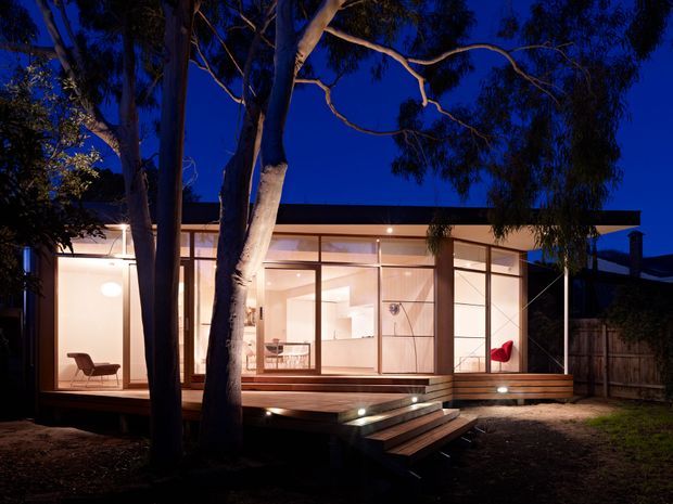 A low, single-storey modern pavilion-like home viewed at dusk through mature trees. The building has extensive floor-to-ceiling glazing, slender dark frames, warm timber decking and steps, and a simple flat roof. Warm interior lighting reveals an open living area with a lounge chair and dining space, while the surrounding garden and trees frame the facade.