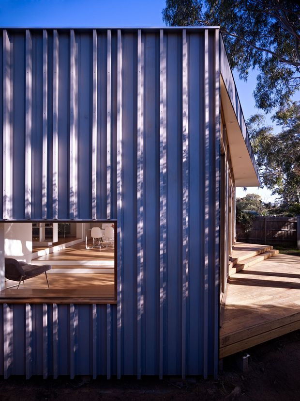 Exterior view of a compact contemporary building clad in vertically ribbed dark blue-grey metal panels. A large window reveals a warm timber-floored interior with modern seating, while a timber deck and steps extend along the side. The simple flat roof, strong vertical facade rhythm, and connection to the landscaped outdoor area create a crisp modern architectural composition.