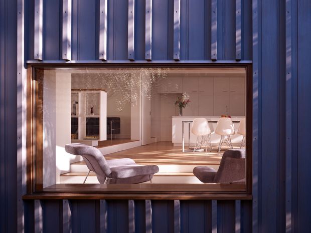 Exterior view through a large rectangular window into a bright contemporary open-plan interior. The building facade is clad in dark blue-grey vertically ribbed metal panels with contrasting timber window trim. Inside, a white kitchen with island seating is visible beyond a warm timber floor, along with sculptural lounge chairs, a fireplace or media wall, and soft pendant or decorative lighting. The image strongly emphasizes the contrast between the dark corrugated exterior envelope and the light, refined interior.