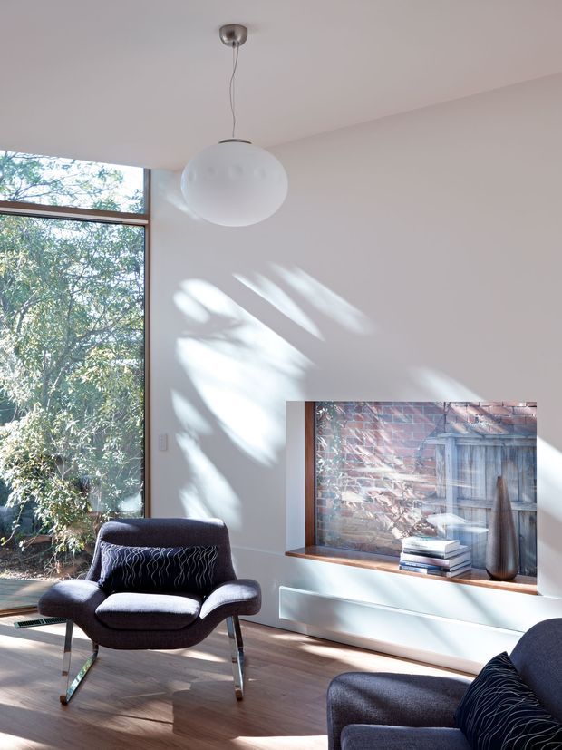 A bright, minimalist living room with white walls, timber flooring, and a large floor-to-ceiling window overlooking leafy greenery. Two dark upholstered lounge chairs sit beneath a simple white pendant light. Sunlight casts strong leafy shadows across the wall, while a low white built-in media console displays a framed artwork, books, and a decorative vase.