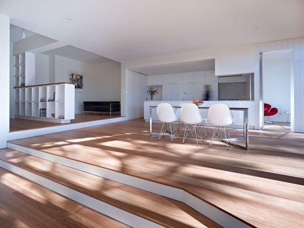 A bright, open-plan contemporary interior with a white kitchen and central island, four white dining stools, integrated cabinetry, and a sunken living area separated by broad timber steps. Built-in white shelving and a dark sofa are visible to the left, while strong sunlight creates long shadows across the timber flooring.