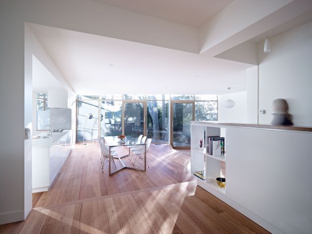 Bright open-plan contemporary interior with warm timber flooring, white walls and built-in cabinetry. The foreground includes a low white partition and open shelving, while a dining table and chairs sit beside extensive floor-to-ceiling glazing. A compact kitchen area is visible to the left, and the room opens toward a sunlit outdoor setting.