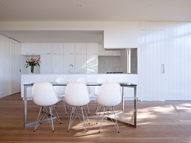 Bright, minimalist open-plan kitchen and dining area with white flat-panel cabinetry, a central island, and a long dining table with six white molded chairs. Warm timber flooring contrasts with the predominantly white joinery and walls. A tiled splashback, integrated appliances, pendant-free recessed lighting, and vertical white wall panelling are visible, with sunlight casting shadows across the floor.