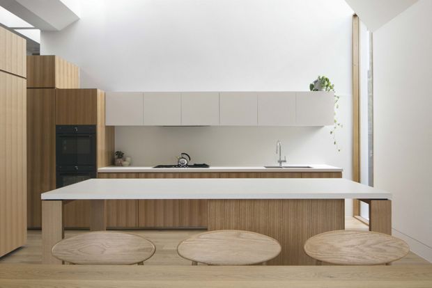 Bright, minimalist contemporary kitchen with a large island and three round timber stools in the foreground. The kitchen features warm vertical timber cabinetry, matte white overhead cabinets, a pale stone island benchtop, integrated appliances, a long horizontal window, and a high white ceiling with a skylight-like opening. The restrained palette and abundant natural light create a calm, clean atmosphere.