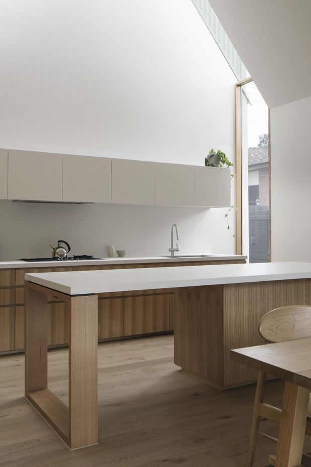 A bright, minimalist kitchen with warm timber cabinetry, a white island benchtop, pale cabinetry, integrated cooking and sink zones, and light timber flooring. A tall glazed opening brings in natural light and overlooks a neighbouring building. A timber dining table and chair are partially visible in the foreground.