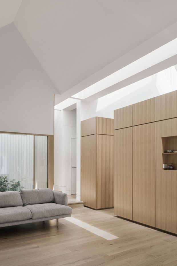 Bright contemporary interior featuring a grey upholstered sofa, pale timber flooring, and extensive floor-to-ceiling light timber cabinetry with integrated shelving. White walls and a sculptural angled ceiling are illuminated by a long skylight or clerestory opening, creating a calm, minimalist living space.