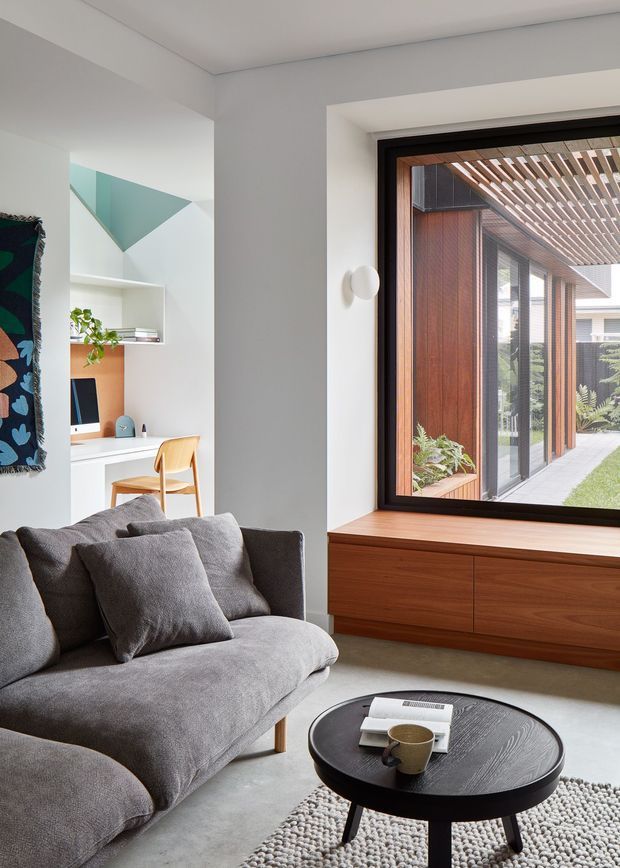 A bright contemporary living room with a large grey upholstered sofa, round dark coffee table, and textured light rug. A built-in walnut timber window seat and cabinetry frame a large dark-framed window overlooking a covered timber courtyard walkway. An integrated study nook with white shelving, a timber desk surface, and a wooden chair is visible to the side, along with a colourful wall textile and a small plant. The room has clean white walls, a simple ceiling, and strong indoor-outdoor connection through the expansive glazing.