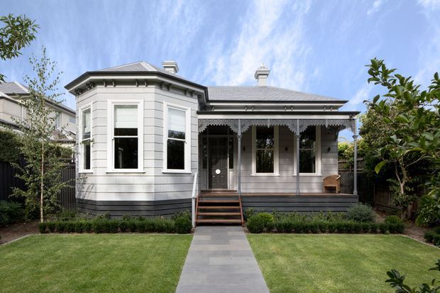 Front exterior view of a restored single-storey timber villa with a projecting bay window, decorative lacework verandah, corrugated metal hipped roof, painted weatherboard cladding, and a symmetrical garden approach. The facade is predominantly light grey and white with dark trim and a central front staircase.