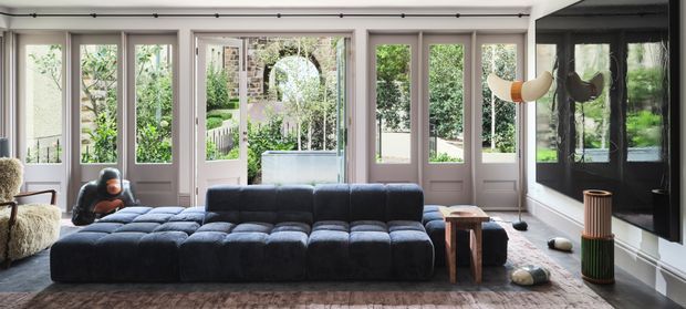 A spacious, light-filled living room with a large modular dark blue sectional sofa, pale walls, extensive timber-framed glazed doors and windows opening toward a leafy garden, and a dark feature wall with sculptural lighting and decorative objects. The room has a refined contemporary character with a warm, eclectic mix of furnishings and finishes.