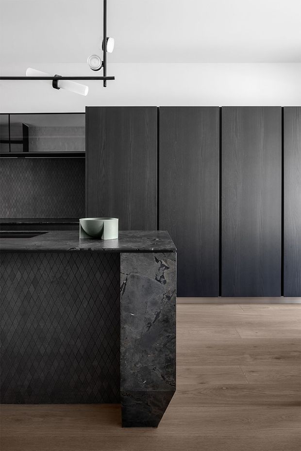 A dark, refined kitchen interior featuring a black textured island with a dramatic natural stone waterfall end, tall dark timber-grain cabinetry, pale timber flooring, and a minimalist linear pendant light. The island front has a diamond-patterned textured finish, while the monochromatic palette is softened by the warm wood floor and small ceramic vessels.