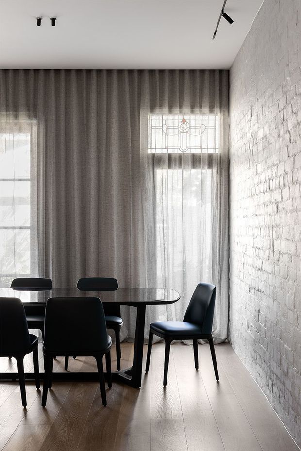 A refined dining area with a dark oval dining table and black upholstered chairs set against a white-painted exposed brick wall. Full-height sheer grey curtains cover tall windows, including a decorative leadlight-style upper window, while pale timber flooring and minimalist track lighting create a bright contemporary interior.
