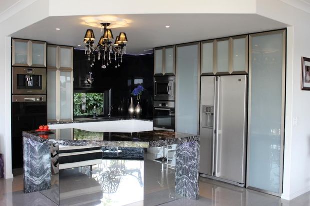 A luxurious contemporary kitchen with a large marble island and waterfall ends, glossy dark cabinetry, integrated stainless-steel appliances, glass-front upper cabinets, and a dramatic crystal chandelier. The open-plan space has polished reflective flooring and a monochromatic black, white, and grey palette.