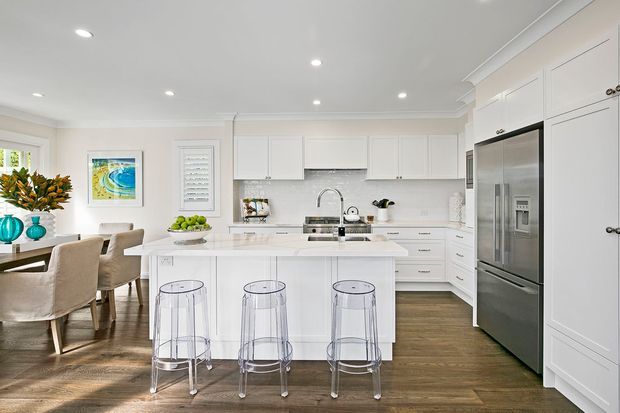 Bright, spacious white kitchen with a large central island, integrated cabinetry, stainless-steel refrigerator, white subway-tile splashback, timber-look flooring, and three transparent bar stools. A dining area with upholstered chairs is visible to the left, while recessed ceiling lights and polished white joinery create a clean, refined interior.