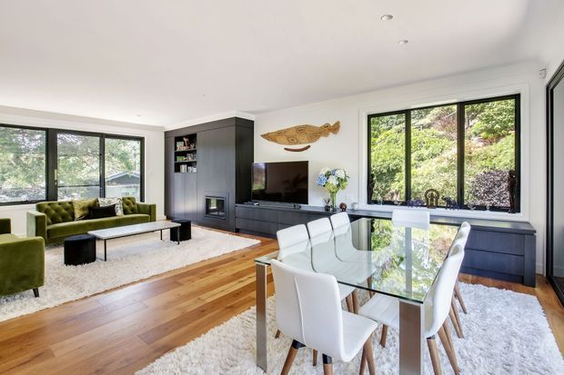 Bright open-plan living and dining room with large black-framed windows, white walls and ceiling, warm timber flooring, a dark built-in media wall with fireplace and shelving, and a glass dining table with white upholstered chairs. A green velvet sofa and armchairs sit over a large pale rug in the living area, with greenery visible outside.