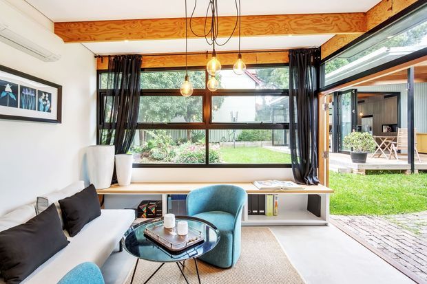 Bright, compact living room with a white built-in window seat and low shelving, white sofa, teal armchairs, round glass coffee table, and exposed timber ceiling beam. Large dark-framed windows and an open glazed door connect the room to a green courtyard and covered outdoor area. Pendant lights, black curtains, and framed artwork add contrast.