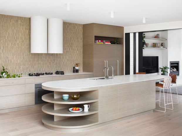 A bright, contemporary open-plan kitchen dominated by a large light-wood island with a white stone benchtop and rounded open shelving at one end. The kitchen features pale wood cabinetry, a textured beige tiled splashback, integrated appliances, white cylindrical rangehoods, and a black-framed opening to an adjacent living area with shelving, television and fireplace visible beyond.