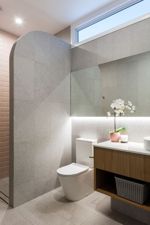 Contemporary bathroom featuring large-format light grey stone-look wall and floor tiles, a curved tiled privacy partition, wall-hung timber vanity with white countertop, integrated under-mirror lighting, frameless mirror, white toilet, and a high horizontal window. A pink-tiled wall provides a subtle contrasting accent.