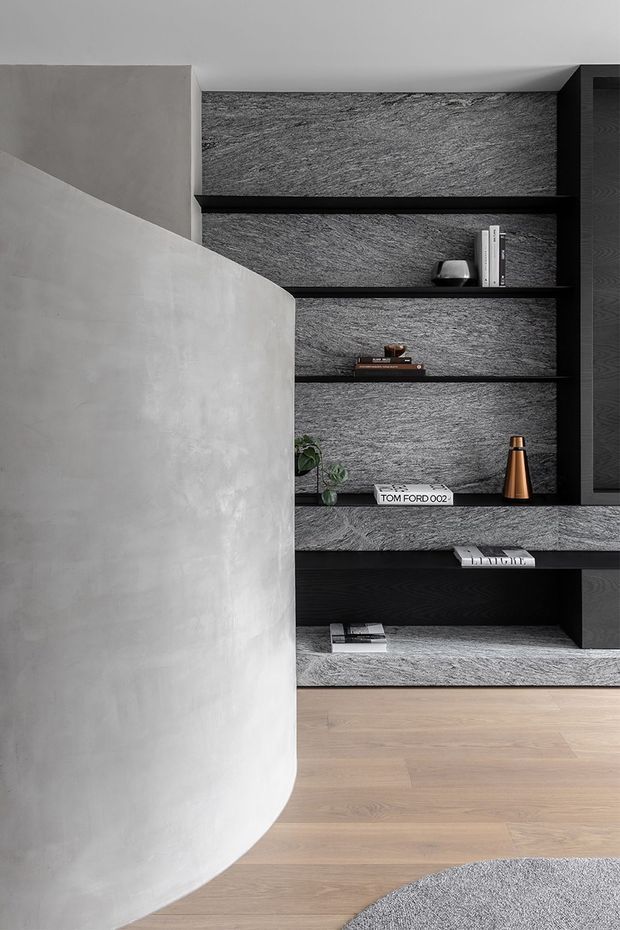 A refined contemporary interior detail featuring a curved light-grey plaster or microcement partition in the foreground and a full-height black built-in shelving wall behind it. The shelving is backed with textured grey stone-like surfacing and styled with books, a small plant, and decorative objects. Pale timber flooring and a grey rug complete the restrained monochromatic palette.