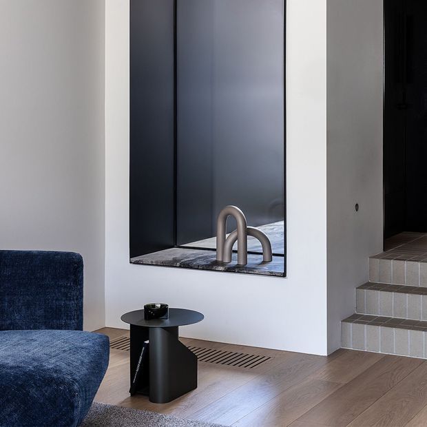A refined contemporary interior corner featuring a deep blue upholstered lounge chair and black sculptural side table beside a large dark-framed mirror. The mirror reflects curved metallic tapware and a tiled wet area, while a short flight of tiled steps leads to a darker adjoining space. Light timber flooring, white walls, and linear floor grilles create a clean, minimalist composition.