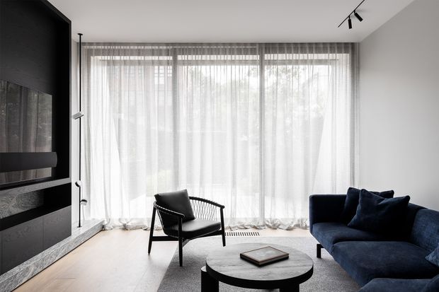 Contemporary living room with a dark built-in media wall, pale timber flooring, a blue upholstered sofa, black lounge chair, round coffee table, and full-height sheer curtains across generous glazing. The room has a restrained monochrome palette with deep navy accents and soft natural light.