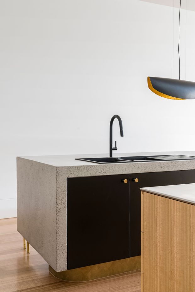 Contemporary minimalist kitchen featuring a large terrazzo-like concrete island with integrated black sink and matte black gooseneck tap. Dark cabinetry with small brass-gold knobs contrasts with a light timber island, pale walls, timber flooring, and a sculptural black-and-gold pendant light.