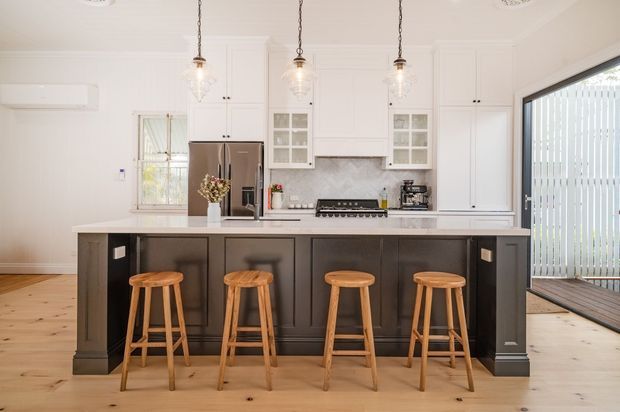 Bright, spacious contemporary kitchen with a large dark island and four timber stools, white shaker-style cabinetry, marble-look splashback, stainless-steel refrigerator, pendant lights, and pale timber flooring. A glazed opening connects to an outdoor deck or balcony.
