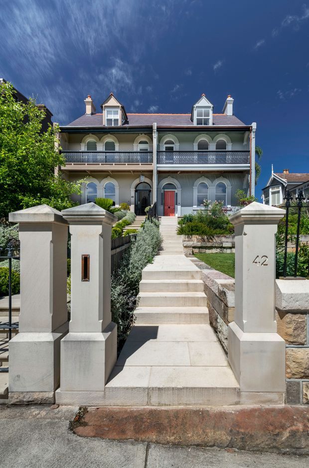 Street-facing view of a grand heritage terrace or townhouse with a symmetrical pale rendered facade, ornate arched windows, decorative iron balcony balustrades, dormer windows, chimneys, and a red front door. A formal landscaped entry garden is approached by a broad flight of pale stone steps between substantial rendered gate pillars marked 42. The image is photographed in bright sunlight against a deep blue sky.