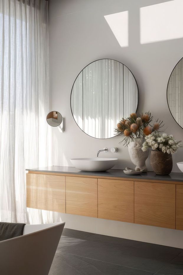 A refined contemporary bathroom with a floating timber vanity, dark stone-look benchtop, white vessel basin, and round wall mirror. Sheer floor-to-ceiling curtains filter abundant natural light, while a freestanding bathtub is partially visible in the foreground. Decorative dried floral arrangements add organic texture to the calm, neutral space.
