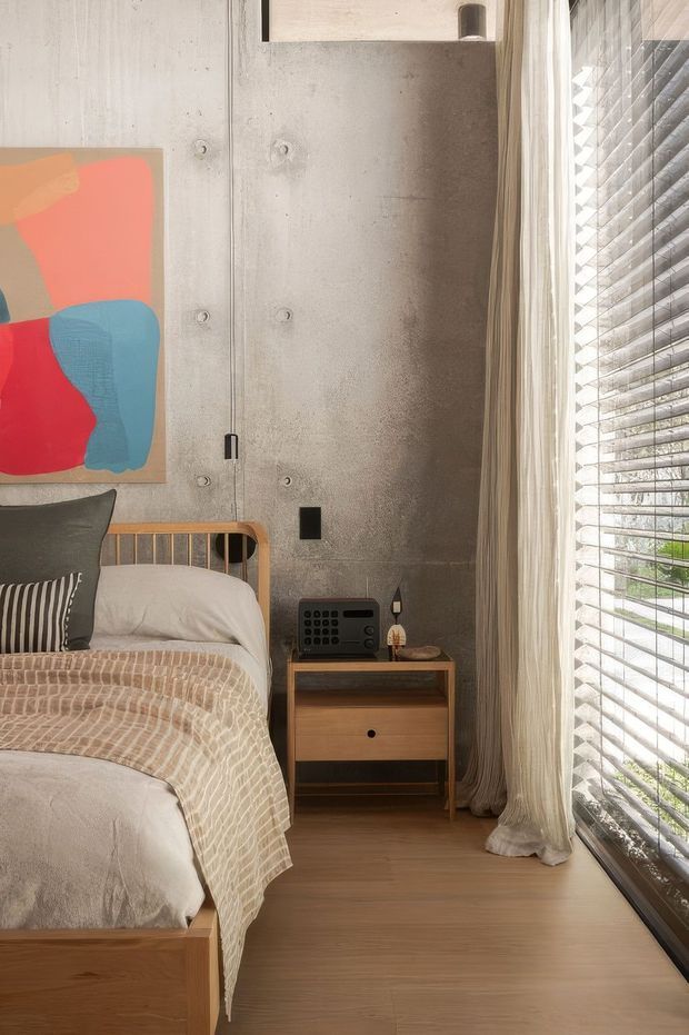 A warmly lit contemporary bedroom with a partially visible timber bed, neutral bedding, a colourful abstract artwork, and a compact mid-century-style timber bedside table. The room features a raw concrete wall with visible form-tie marks, timber flooring, full-height curtains, and a large window fitted with horizontal blinds.