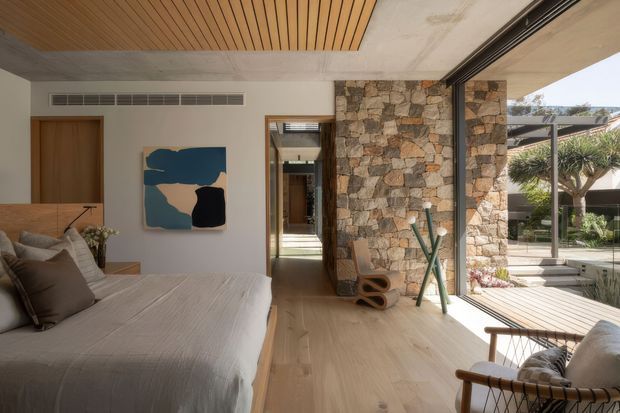 A contemporary bedroom with a low platform bed, neutral linen bedding, timber flooring, and a large glazed opening to a landscaped outdoor area. A rustic stacked-stone feature wall frames the hallway, while a slatted timber ceiling adds warmth above the room. The space combines natural materials, restrained furnishings, and abundant daylight.