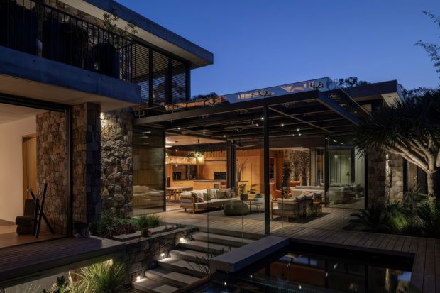 Twilight exterior view of a contemporary multi-level residence with extensive floor-to-ceiling glazing opening onto a covered outdoor living area. The home features dark metal framing, stone-clad walls, timber decking, a rooftop or pergola-like structure, landscaped planting, and a reflective swimming pool in the foreground. Warm interior lighting reveals an open-plan living and kitchen area.