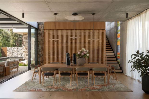 A refined open-plan dining area in a contemporary home, centered on a long timber dining table with upholstered chairs and a patterned rug. A full-height vertical timber batten screen conceals the stairway and forms a warm feature wall, while exposed stonework, concrete ceilings, extensive glazing, and sheer curtains create a connection to the landscaped exterior. The space has a calm, natural, sophisticated atmosphere.
