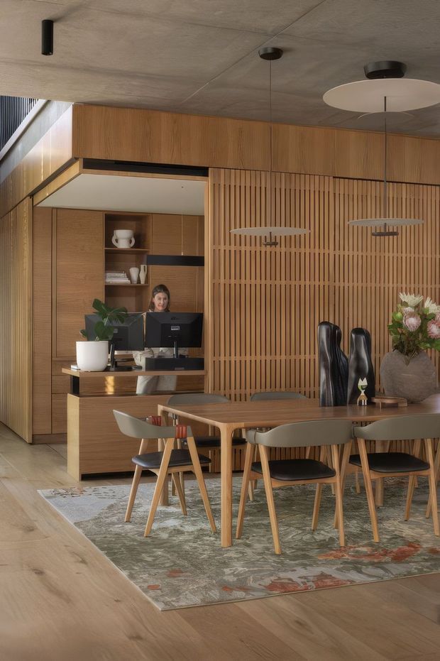 A warm, contemporary dining and reception area with extensive timber joinery. A rectangular timber dining table is surrounded by upholstered timber-framed chairs on a patterned rug, while a built-in reception or service counter with computer monitors occupies the rear. Vertical timber battens form a feature partition, complemented by a concrete-look ceiling, integrated cabinetry, pendant lighting and decorative objects.