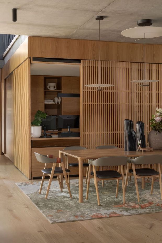 Warm contemporary dining and kitchen interior with extensive timber cabinetry and vertical timber slat screening. A rectangular timber dining table is surrounded by upholstered dining chairs over a muted patterned rug. Behind it, a compact dark kitchen with integrated appliances and open shelving is partially concealed by the slatted partition. The space has a smooth concrete-look ceiling, minimalist pendant lighting, pale timber flooring, and restrained natural decor.