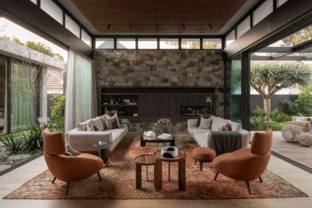 A refined living room with expansive floor-to-ceiling glazing, clerestory windows, a warm slatted timber ceiling, and a large textured stone fireplace wall with integrated dark joinery. Two pale sofas and sculptural rust-toned lounge chairs surround layered coffee tables and a patterned rug, while landscaped garden areas are visible through the open glazing.