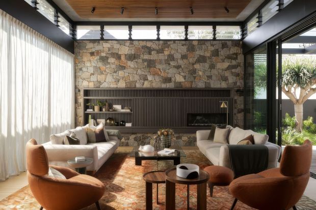 A sophisticated living room with two cream sofas and sculptural rust-orange lounge chairs arranged around nested coffee tables. The room features a full-height irregular natural-stone feature wall with dark vertical timber-look built-in media joinery, a warm slatted timber ceiling, clerestory glazing, black-framed full-height glass doors, sheer curtains, and abundant garden views. The overall aesthetic is warm, refined, and contemporary with strong indoor-outdoor connection.
