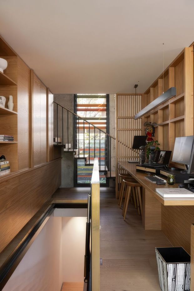 A narrow, warm-toned home office or study arranged along a stairwell. Floor-to-ceiling timber built-in shelving and cabinetry line both sides, with a long integrated desk, stools, computer monitors, books and decorative objects. A black metal stair balustrade and glazed rear opening bring light into the compact split-level space. The design features strong vertical timber slats, dark timber flooring, and a restrained contemporary built-in fit-out.